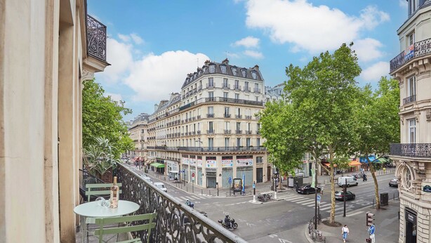 Un balcon avec une table et des chaises, donnant sur une rue bordée d’immeubles et d’arbres.