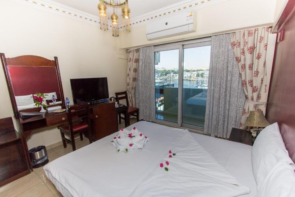Habitación de hotel con cama, escritorio con computadora, televisor y vista a un paisaje urbano a través de un ventanal amplio.