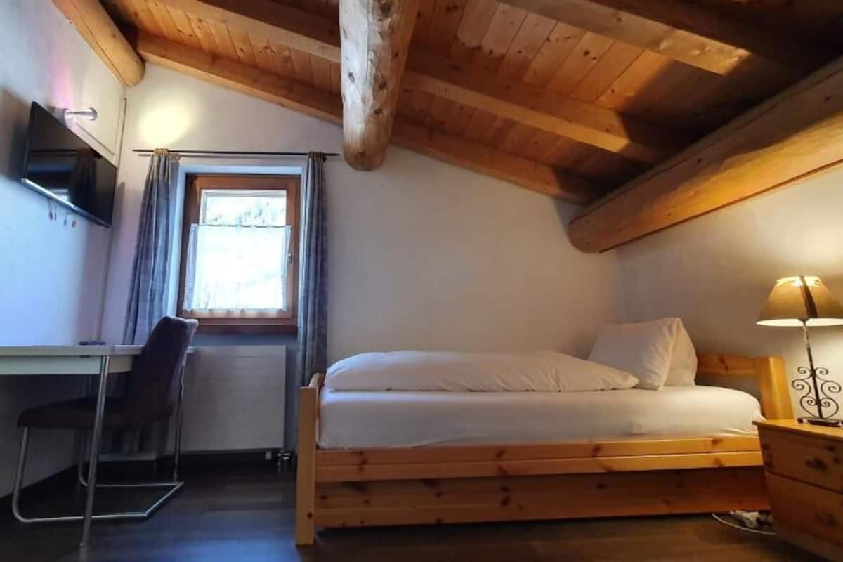 A bedroom with a wooden bed, a desk with a chair, a TV mounted on the wall, and a window with curtains.