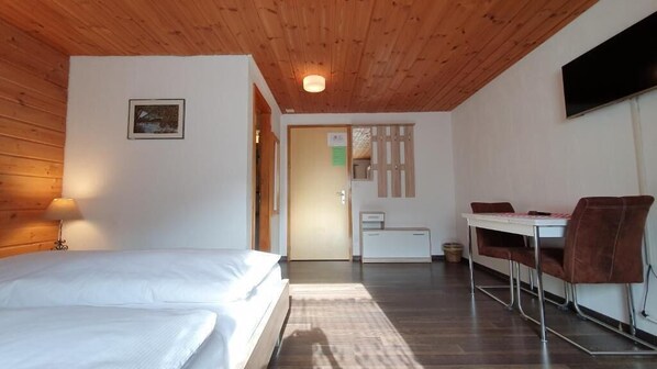 A bedroom with a wooden ceiling, a bed, a desk with a chair, and a television on the wall. - Hotel Suretta (Rheinwald)