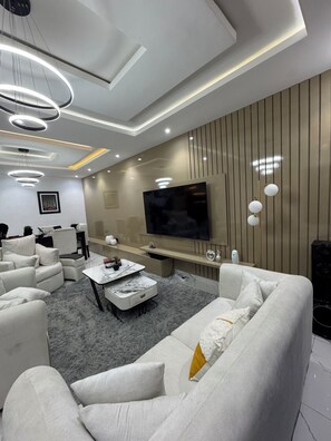 A modern living room with a large flat-screen TV, white sofas, a marble coffee table, and a grey shaggy rug. - Luxury 3-Bedroom Haven in Lekki with 24/7 Power, High-Speed Wi-Fi & Private SUV (Lagos)