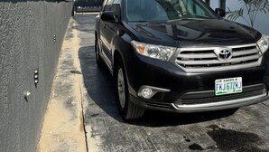 A black Toyota parked in a narrow alleyway between modern buildings with balconies and a clear blue sky.
