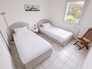 A bedroom with two beds, a small table with a lamp, a chair, and a window with a view of greenery. - Family Apartment with Air Conditioning and Parking in Collioure (Collioure)