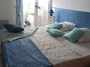 Ein Bett mit gemusterten Betttextilien, ein Nachttisch mit Lampe und ein Fenster mit Vorhängen.