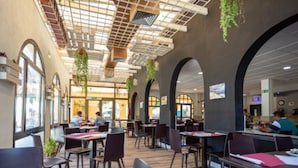 Un restaurant avec un plancher, des tables et des chaises en bois. De grandes fenêtres et des plantes suspendues décorent l’espace.