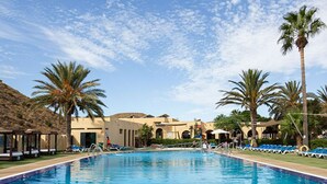 Une piscine entourée de chaises longues et de palmiers, avec des bâtiments aux arches et un ciel dégagé.