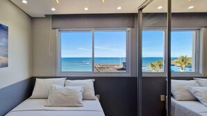 Ein Schlafzimmer mit einem Bett, weißen Kissen und einem großen Fenster mit Blick auf den Ozean und einen Strand.
