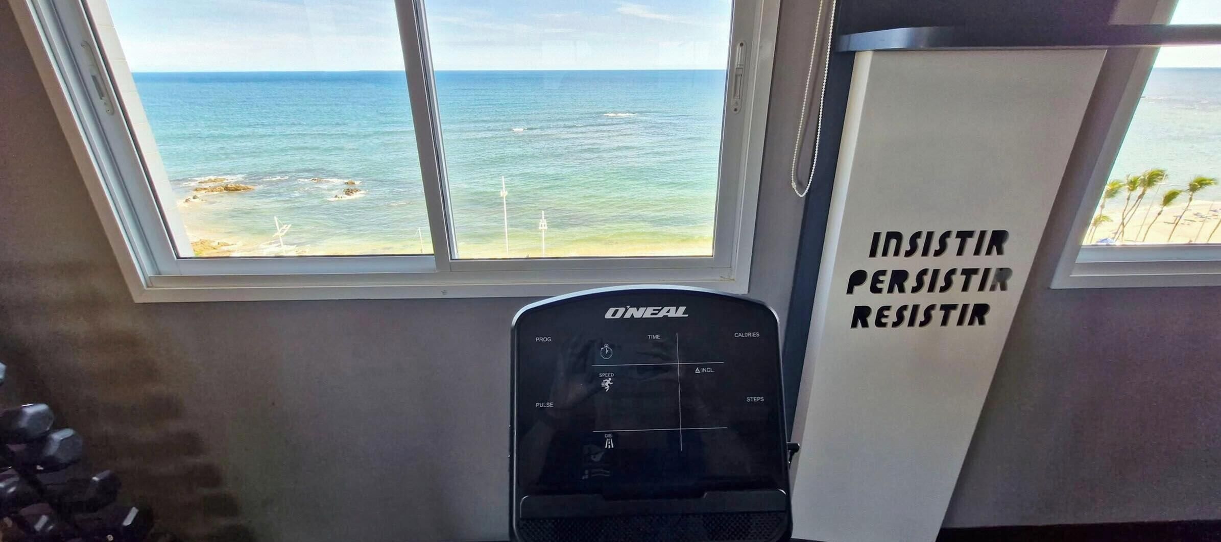 A gym with a view of the ocean through a window, a treadmill, and a motivational poster.