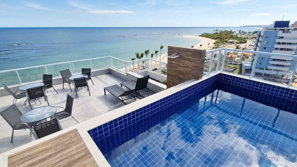Ein Pool auf dem Dach mit Blick auf das Meer und den Strand, umgeben von Sitzgelegenheiten im Freien und einem modernen Gebäude im Hintergrund.
