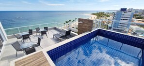 A rooftop pool with a view of the ocean and beach, surrounded by outdoor seating and a modern building in the background. - COPIA0105 - Edifício Essencial Piata (Salvador)