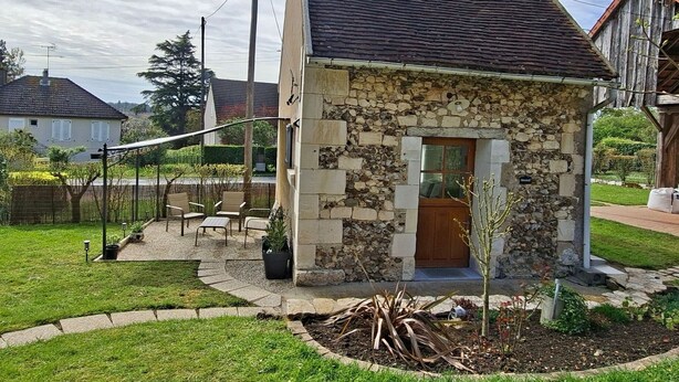 Une maison en pierre avec une porte en bois, un petit jardin et un espace extérieur aménagé pour s’asseoir.