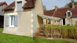 Une maison à deux étages, dotée d’un toit en pente, d’une cheminée et d’un petit jardin planté de jeunes arbres.