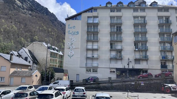 Un hotel de varios pisos con un cartel que dice "Le Lys" y un estacionamiento con varios coches.
