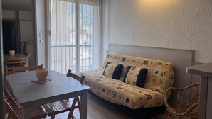 Una sala acogedora con un sofá, una mesa de comedor y una silla. Hay una ventana con cortina, y una lámpara fijada a la pared.