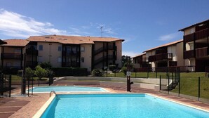 Uma área de piscina sob um céu claro, cercada por edifícios altos com varandas.
