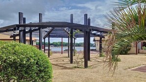 Uma área externa coberta com estrutura de pérgola, um pequeno arbusto e vista para um corpo d'água ao fundo.