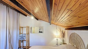 Un dormitorio con techo de madera, una cama con colchón acolchado, un ventilador de pie, un espejo y una lámpara de noche.