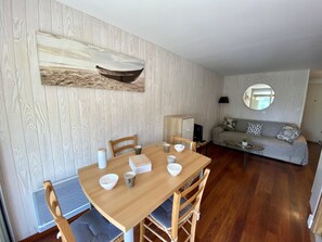 A dining area with a wooden table and chairs, a sofa, a round mirror, and a painting of a boat on the wall. - Apartment for 5 with Garden in Saint Pierre (Fleury-Saint-Pierre-la-Mer)