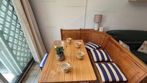 A room with a wooden table set for two, a green sofa with striped cushions, a small plant, and a window with a view of buildings. - 4-Person Apartment with Terrace and Parking in Narbonne (Narbonne)