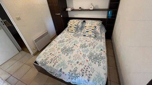 A bedroom with a bed, a wardrobe, a shelf with decorative items, and a wall-mounted light fixture. - 4-Person Apartment with Terrace and Parking in Narbonne (Narbonne)