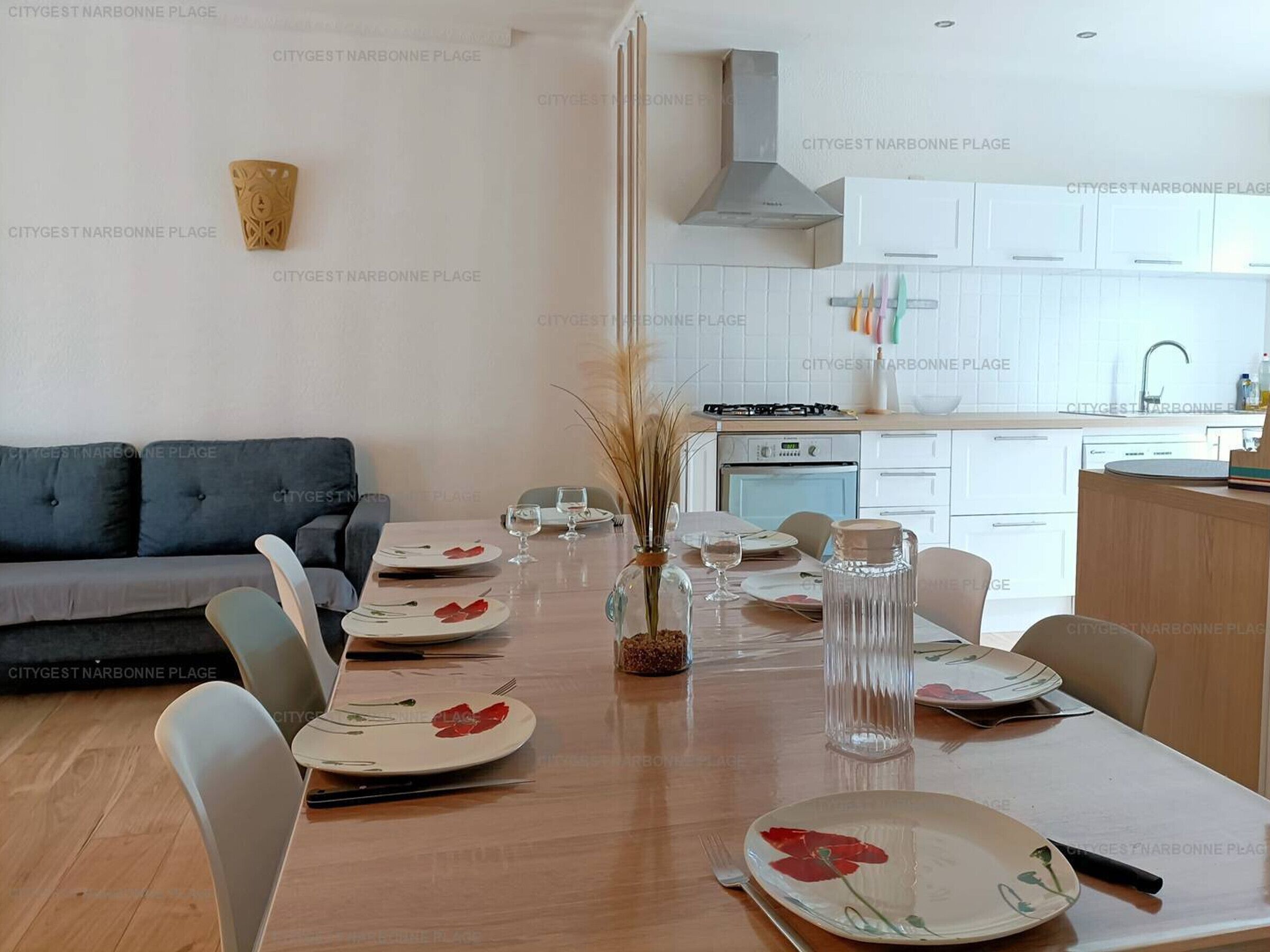 A modern dining area with a long table set for six, a grey sofa, and a kitchen in the background.