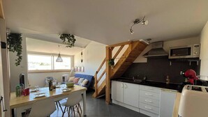 A kitchen with a wooden staircase, white cabinets, a microwave, and a dining area with a table and chairs. - Cozy Nest in Narbonne for 4, near beach, includes parking (Narbonne)