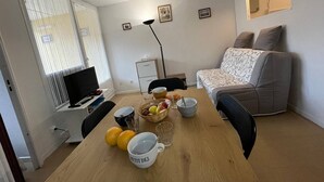 Ein kleiner, gut beleuchteter Raum mit einem Esstisch für zwei Personen, einer Couch, einem Fernseher und einem Fenster mit Jalousien.