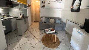Ein kompakter Wohnraum mit einer Küchenzeile, einem grauen Sofa, einem kleinen Rundteppich und einem an der Wand befestigten Spiegel.