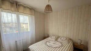 A bedroom with a bed, a window with curtains, a wooden floor, a bedside table, and a hanging light fixture. - 2-Star Apartment 50m from Beach with Balconies and Private Parking (Narbonne)
