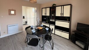 A dining area with a round table, chairs, a hanging light fixture, a cabinet with shelves, and a television. - 2-Star Apartment 50m from Beach with Balconies and Private Parking (Narbonne)