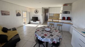 A compact living space with a sofa, a dining table with four chairs, a TV, a kitchen area with a microwave, and a clock on the wall. - Air-Conditioned 3-Room Apartment with Terrace, Close to Beach (Port-la-Nouvelle)