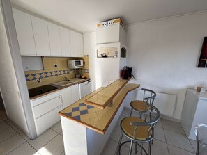 Une cuisine compacte avec des meubles blancs, un four à micro-ondes, un évier et un petit réfrigérateur. On y trouve deux tabourets de bar et un pare-feu en carreaux avec un motif géométrique.
