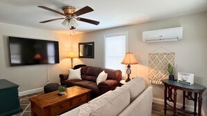 Een woonkamer met een plafondventilator, een flatscreen-tv, een bruine leren bank, een beige zitzak, een houten salontafel en een klein bijzettafeltje met een lamp.