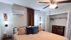Een slaapkamer met een bed, een plafondventilator, een airconditioningapparaat, een nachtkastje, een wandlamp en een kamerbreed tapijt met een patroon.