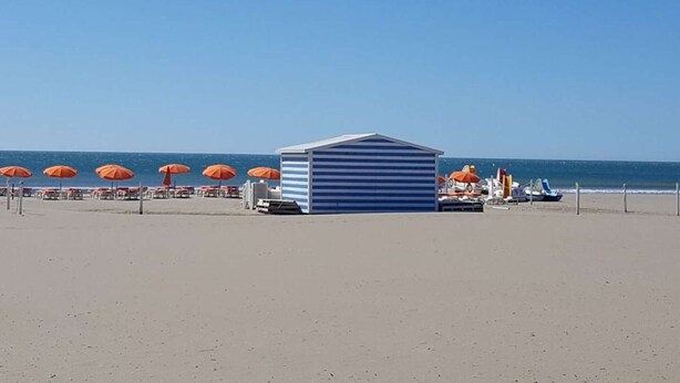 En strand med en randig byggnad, orange parasoller och båtar i bakgrunden.