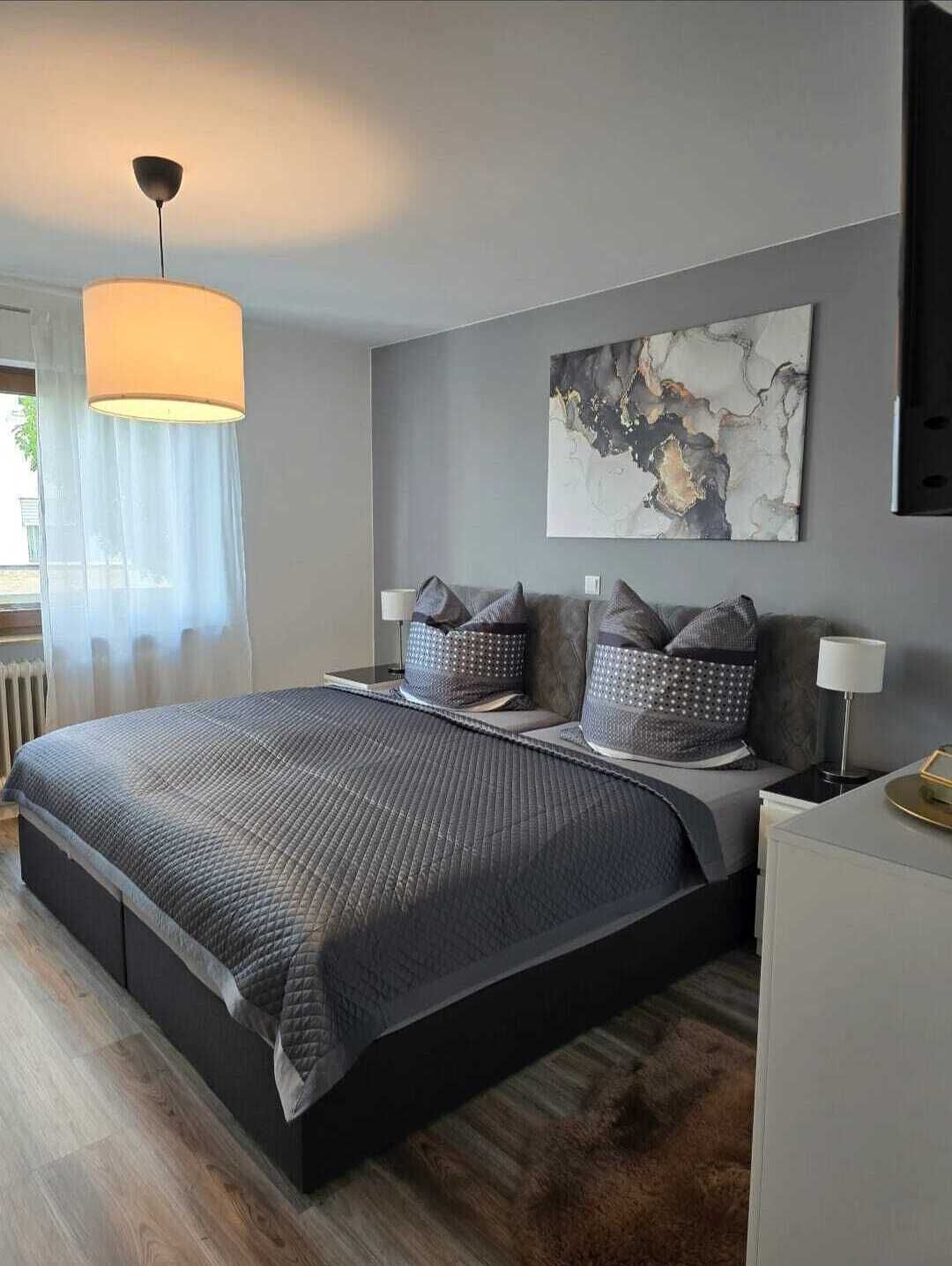 Ein modernes Schlafzimmer mit einem großen Bett, grauen Wänden, einer Deckenleuchte und einem abstrakten Kunstwerk.