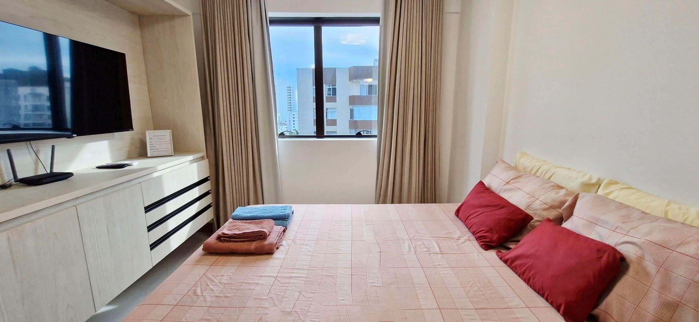 Une chambre d’hôtel avec un lit, un téléviseur fixé au mur, une fenêtre avec des rideaux et une vue sur les bâtiments environnants.