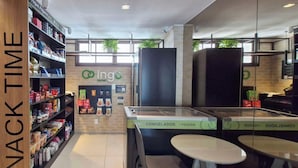 A snack bar area with shelves stocked with various food items, a freezer section, and a seating area with tables. - COBAR0145 - Edifício ZN Barra (Salvador)