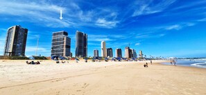 Una playa con edificios altos al fondo, sombrillas y gente.
