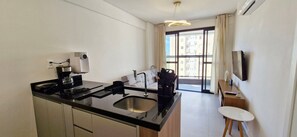 Una cocina moderna con encimera negra, pileta de acero inoxidable y cafetera. Hay una zona de comedor con una mesa y sillas blancas, y un televisor montado en la pared.