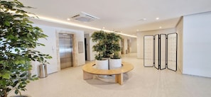 Un moderno vestíbulo de hotel con una mesa redonda de madera, plantas en macetas, un ascensor y un biombo decorativo.