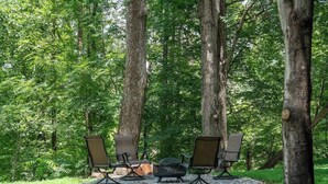 A forest setting with four outdoor chairs arranged around a central fire pit. - Stay Roanoke | Roanoke Roost | 2BR + Fire Pit (Roanoke)