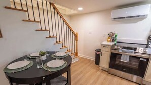 A dining area with a round table set for four, a staircase, and a kitchen with an oven and a trash can. - Stay Roanoke | Roanoke Roost | 2BR + Fire Pit (Roanoke)