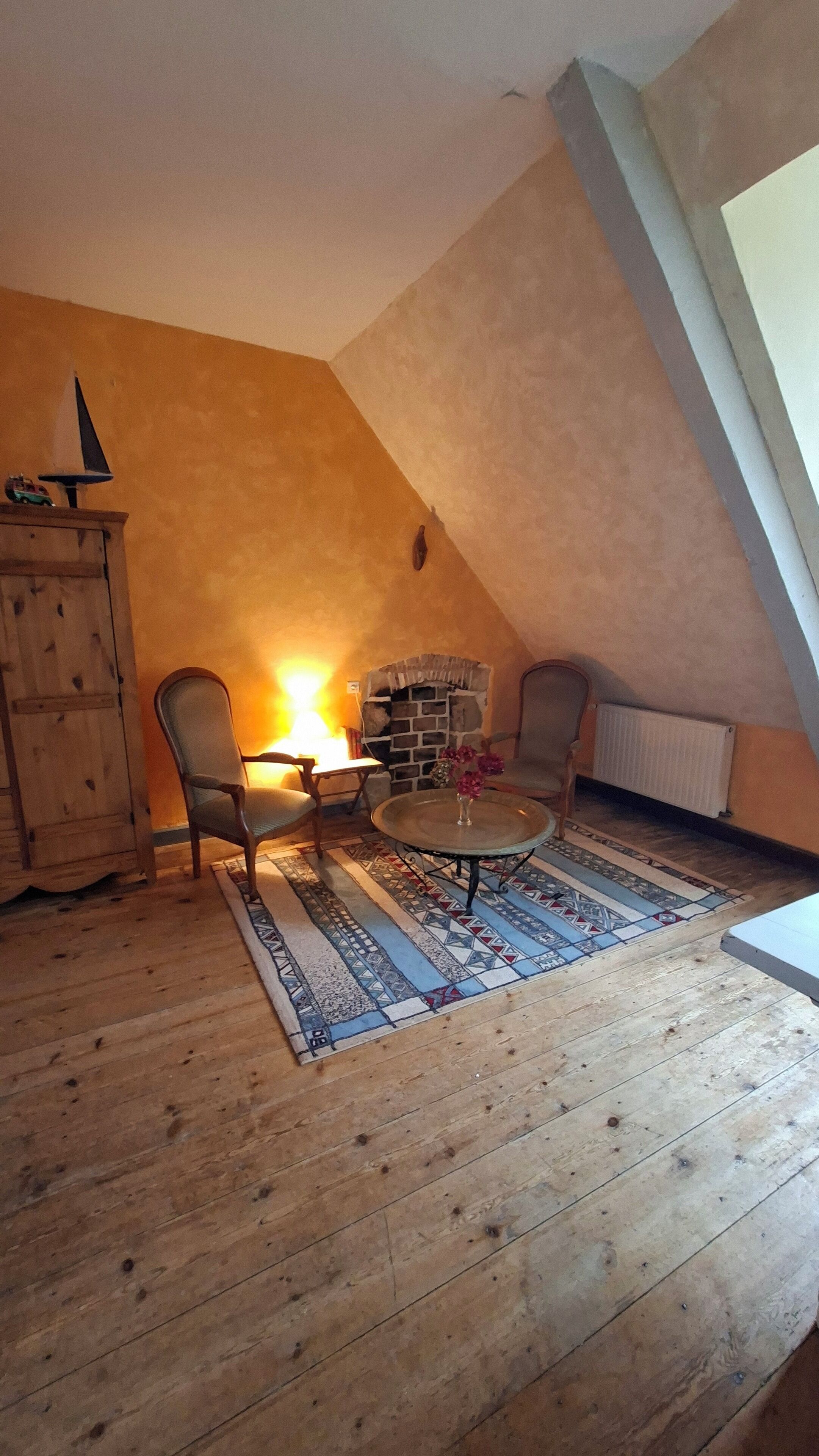 A cozy attic room with a sloped ceiling, a stone fireplace, wooden flooring, and a patterned rug.