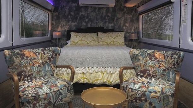 A train compartment with a bed, two patterned armchairs, a small round table, and a window with a view of trees. - Railway train carriage beautifully converted for an unusual stay in Countryside (ELY)
