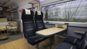 A train carriage with two blue upholstered seats, a small table, and a window with a view of trees. - Railway train carriage beautifully converted for an unusual stay in Countryside (ELY)