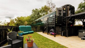 A green camper van parked in a backyard with a wooden fence, a green sofa, and a small table with a potted plant. - Railway train carriage beautifully converted for an unusual stay in Countryside (ELY)