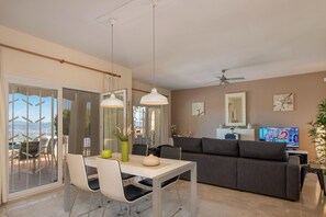 A modern living room with a sofa, a dining table with chairs, a TV, and a balcony with outdoor furniture. - 3BR Apartment With Private Garden (Alhaurin de la Torre)