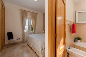 A bedroom with a bed, a chair, a window with curtains, and a bathroom with a sink and mirror. - 3BR Apartment With Private Garden (Alhaurin de la Torre)