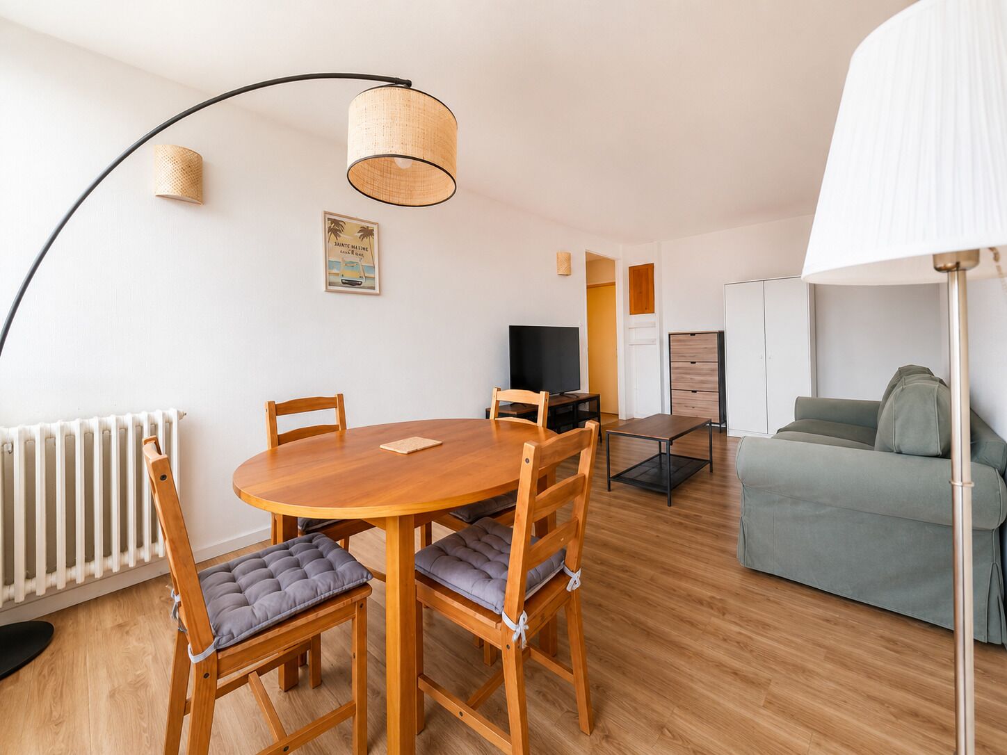 Una sala de estar moderna con una mesa de comedor redonda de madera, dos sillas, un sofá, un televisor y una lámpara de pie.